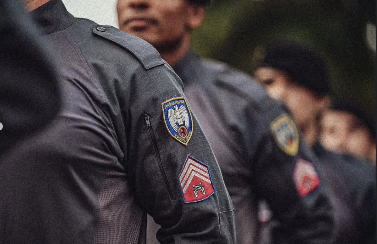 Close da divisa de aluno sargento na manga do uniforme, com a fila do pelotão desfocada ao fundo.